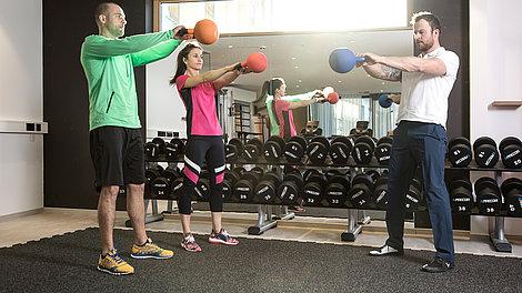 Trainer zeigt zwei Gästen eine Fitnessübung mit Gewicht